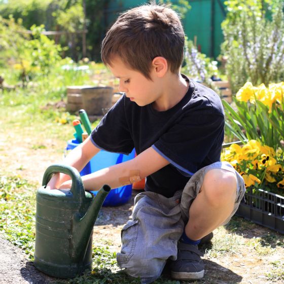 Ein Junge mit kurzen Hosen kniet neben einer dunkelgrünen Gießkanne im Garten der Schule und befeuchtet seine Hände in der Kanne
