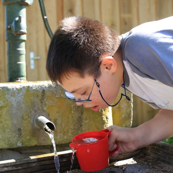 Wasserspiele im Sommer: Ein Junge beugt sich über einen Brunnen und gießt in einem Kännchen Wasser dazu