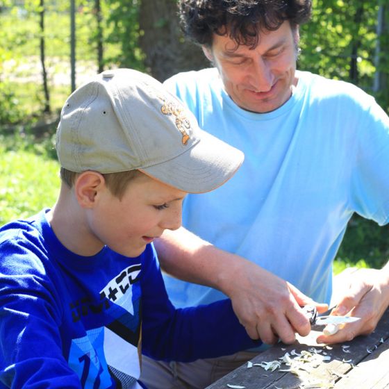 Ein Junge schnitzt mit einem Messer und wird dabei von einem Lehrer begleitet im sommerlichen Garten der Schule