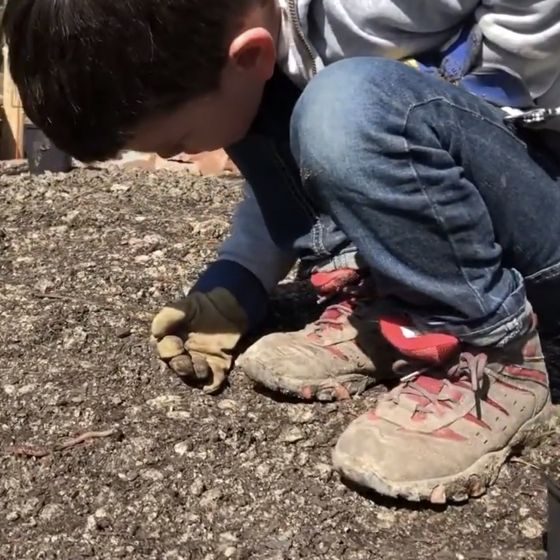 Arbeiten in unserem Garten: ein Junge mit Handschuhen und Arbeitsschuhen kniend und einen Regenwurm betrachtend