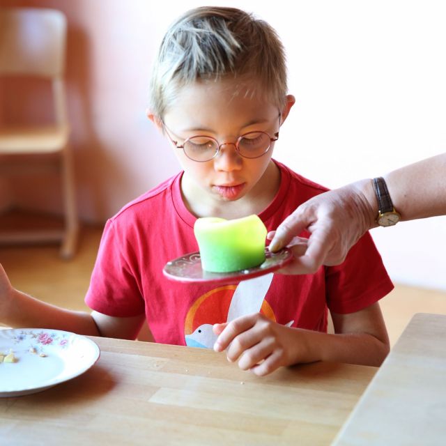 Ein Junge im roten Shirt und Brille bläst eine Kerze aus, die ihm eine Lehrerin reicht
