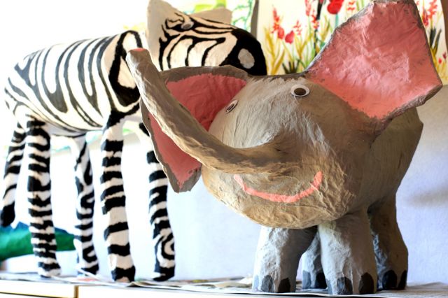 Kunstwerke aus Pappmaché - ein lächelnder Elefant mit rosa Ohren und eine Giraffe im Hintergrund