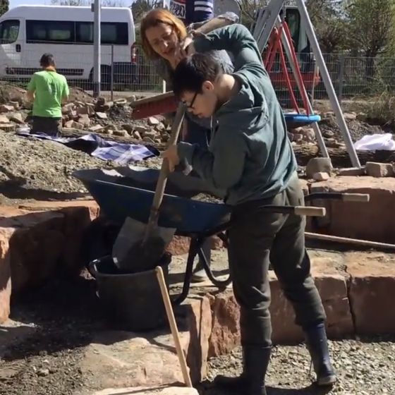 Bauarbeiten in unserem NaturErlebnisRaum: eine Lehrerin hilft einem Jungen mit Schubkarre und Schaufel beim Arbeiten