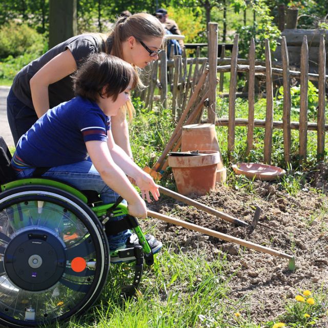 Kind im Rollstuhl arbeitet im Garten mithilfe einer Betreuerin